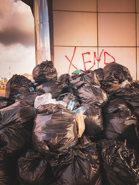 A collection of black rubbish bags piled against a weathered red metal door set into a beige concrete wall. The bags vary in size and are tightly packed, with some partially torn, revealing contents i